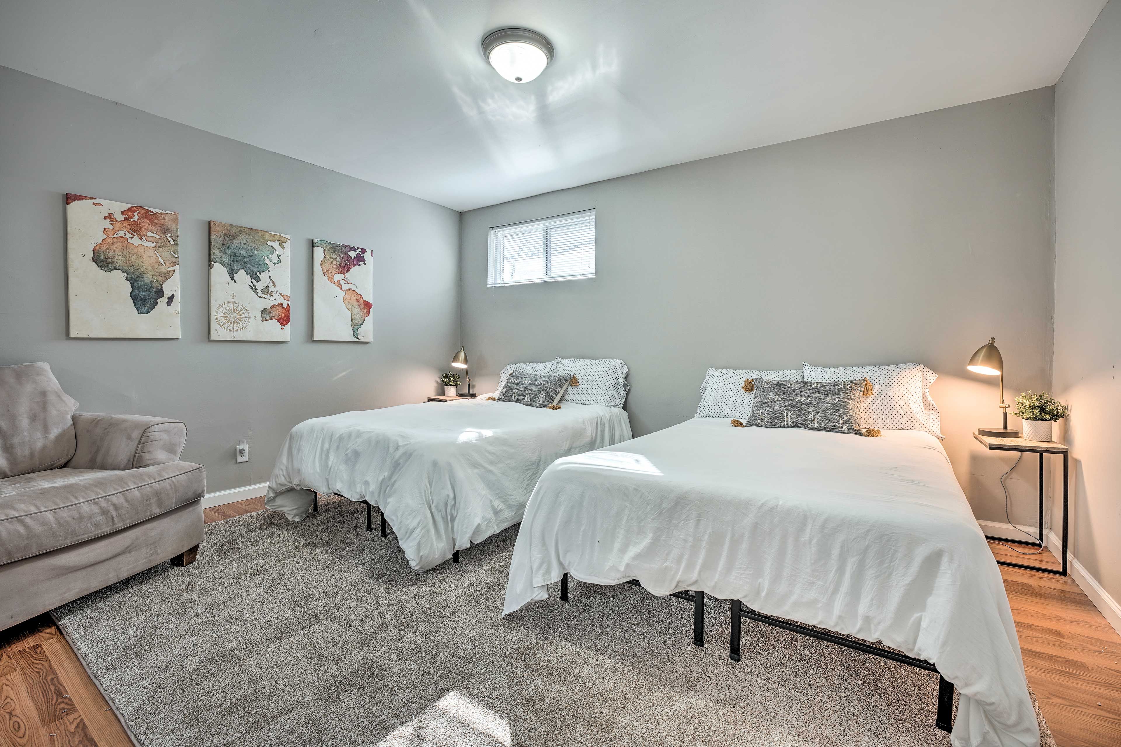 A bedroom with two beds, a grey sofa, a world map artwork on the wall, and a window with blinds.