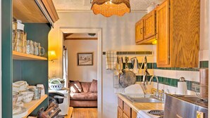 A compact kitchen with wooden cabinets, a stove, and a sink. There are shelves with kitchenware and a dining area visible in the background. - Screened Porch: 'the Rock Cottage' in Hot Springs (Hot Springs)