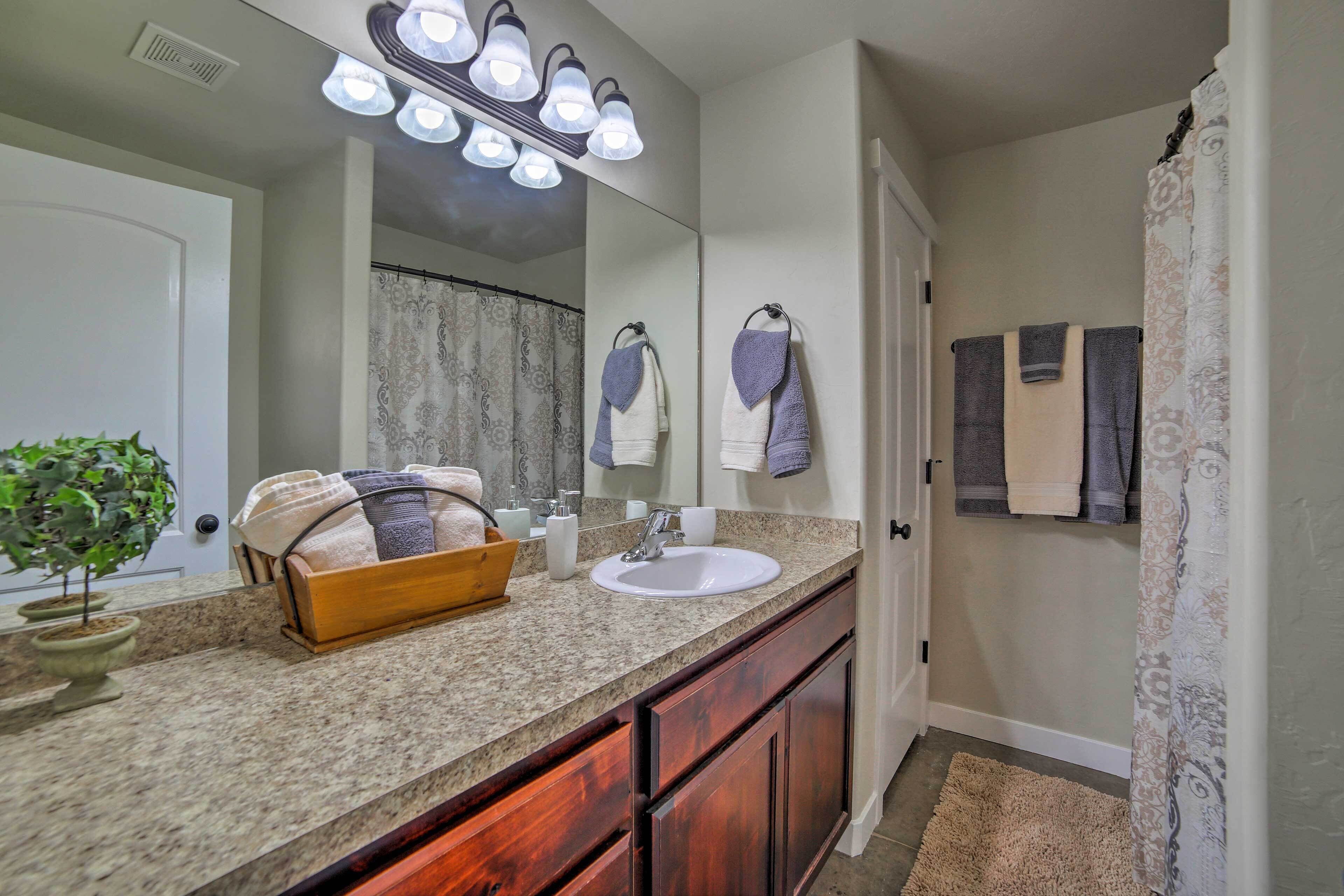 A bathroom with a granite countertop, wooden cabinets, a sink, a mirror with lights, a potted plant, and towels hanging on the wall.