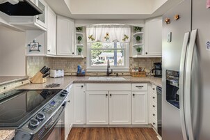 Una cocina con armarios blancos, horno empotrado, refrigerador de acero inoxidable y una ventana con cortina de girasol.