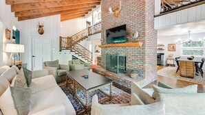 A spacious living room with a brick fireplace, white walls, and a wooden staircase. - Chic Highlands Cabin Retreat w/ Deck + Office! (Highlands)