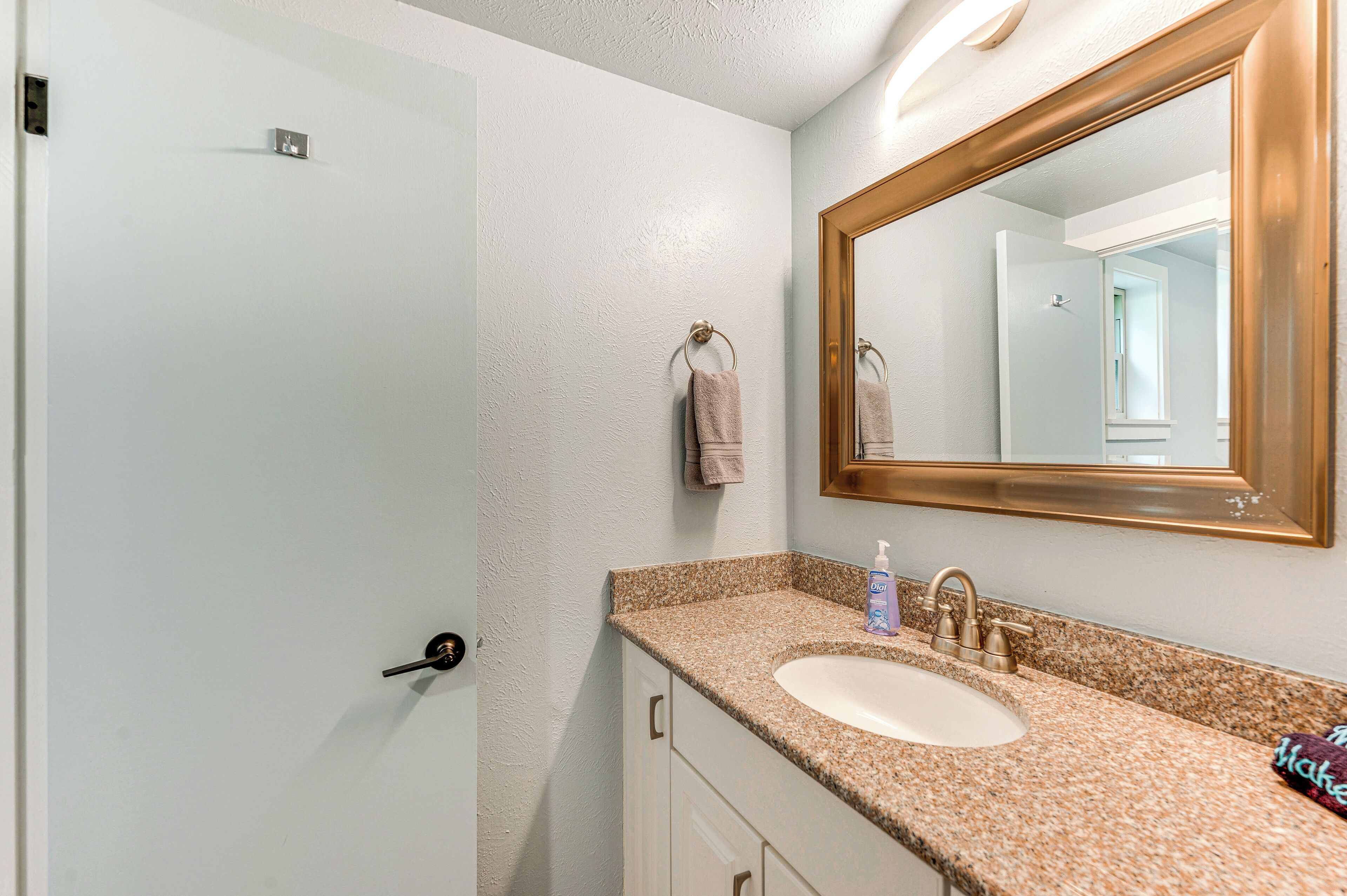 A bathroom with a white door, a sink with a granite countertop, a mirror with a gold frame, and a towel hanging on the wall.