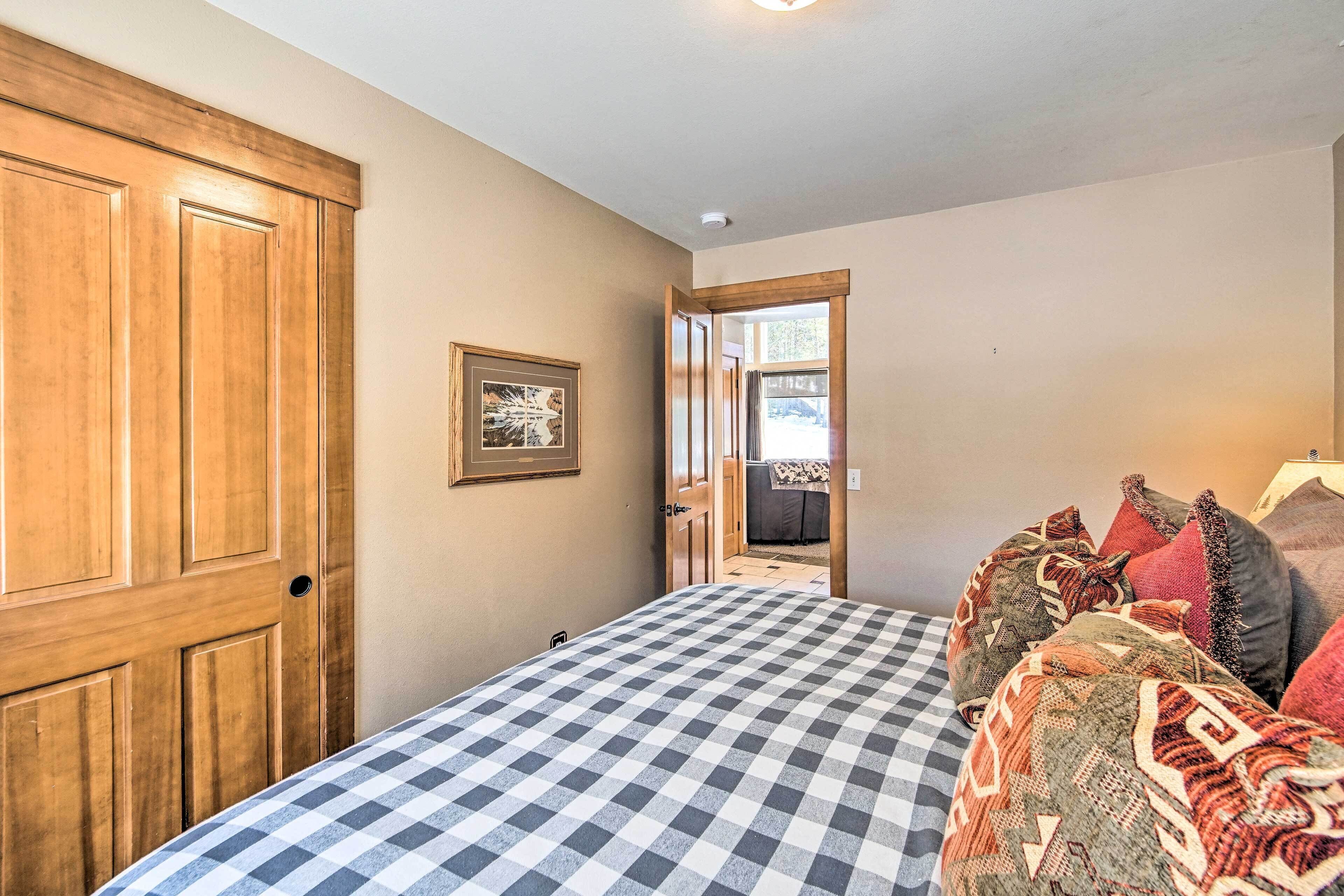 A bedroom with a checkered bedspread, a wooden wardrobe, a framed picture on the wall, and a view into another room through an open door.