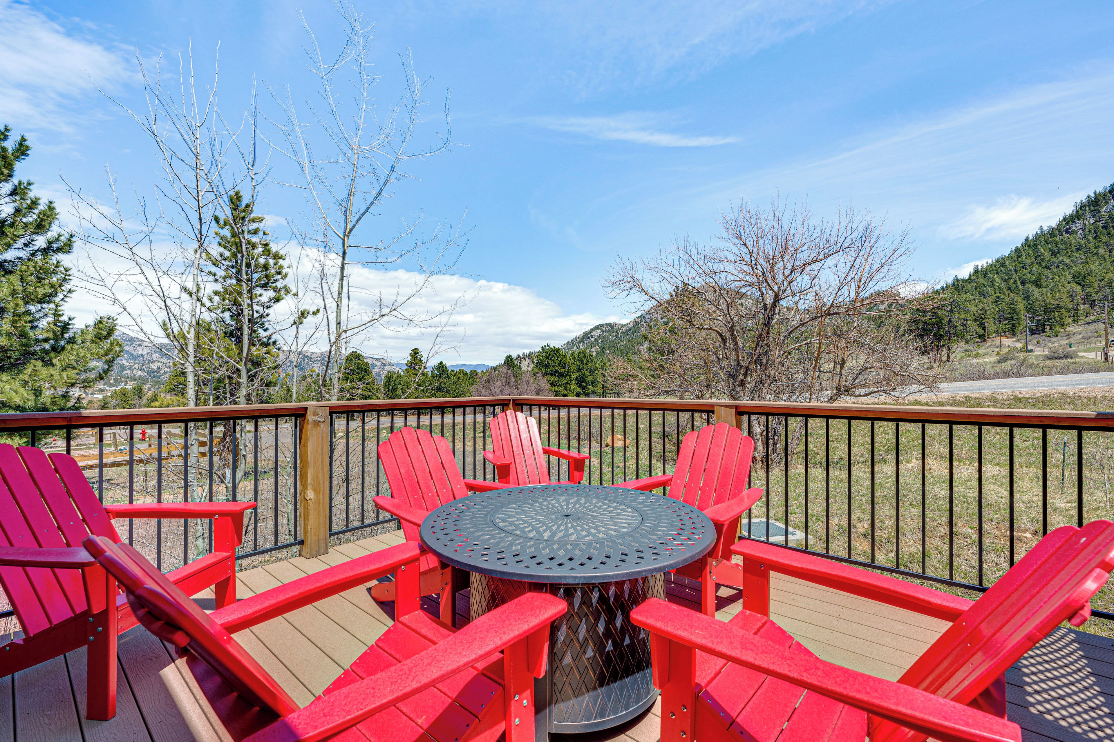 A deck with a round table and six red Adirondack chairs, overlooking a mountainous landscape.