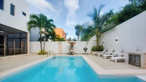 Una piscina con sillas de descanso y una sombrilla, rodeada por un muro blanco y palmeras.