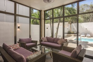 A sunlit room with wicker furniture, a glass table, and a view of a pool and palm trees.