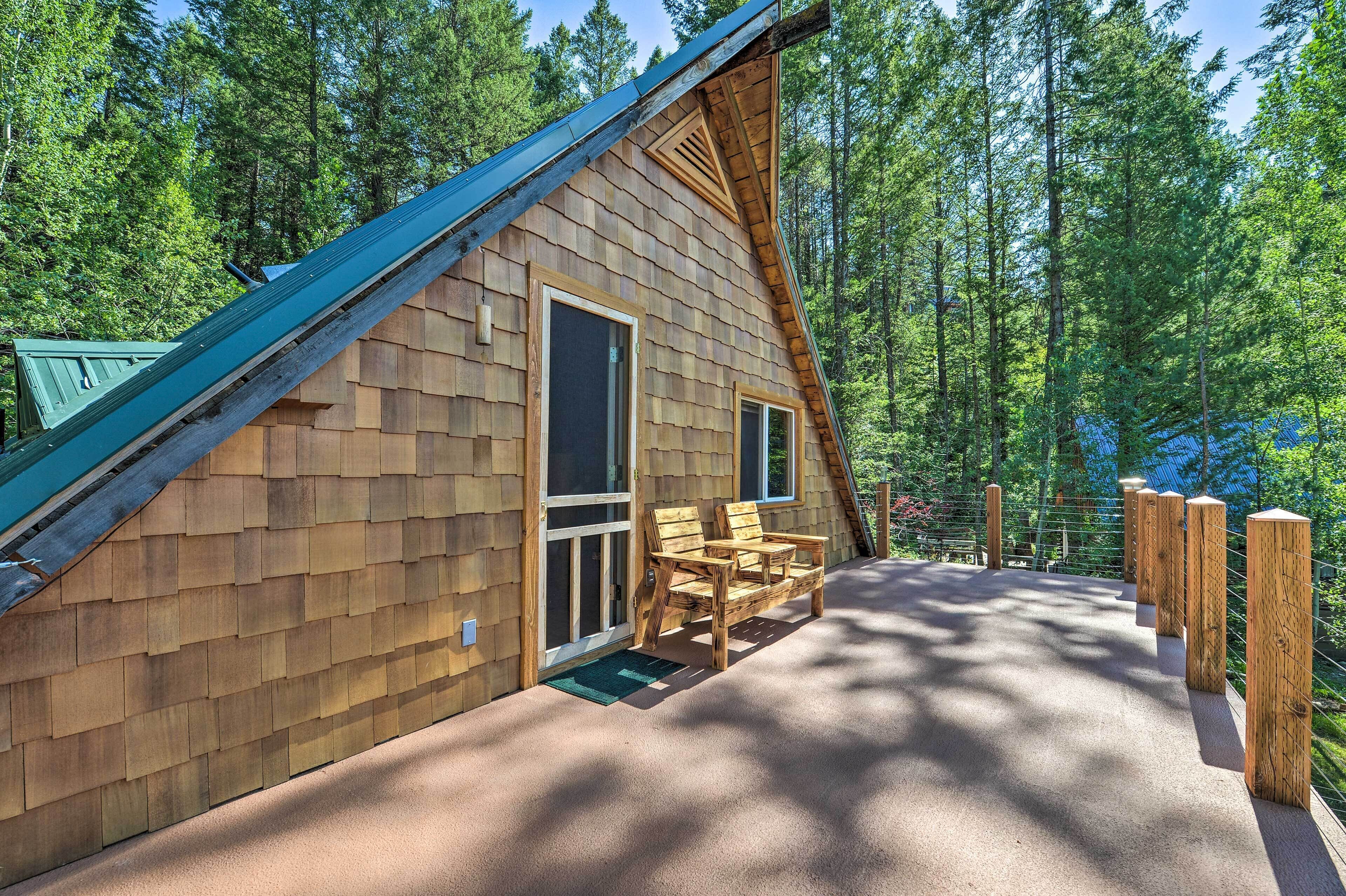 Una cabaña de madera con techo inclinado, un porche cubierto con un banco y un bosque de fondo.