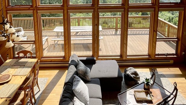 Un intérieur de chalet chaleureux avec un grand plafond en bois, un canapé d’angle, une table à manger et une vue sur une terrasse et les montagnes, le tout visible à travers de grandes fenêtres.