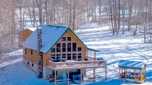 Une cabane en rondins dotée d’une grande véranda et d’un espace extérieur aménagé, le tout entouré d’un paysage enneigé.