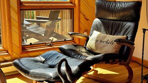 Un fauteuil en cuir avec un coussin, donnant sur une terrasse en bois et des montagnes, vue par une fenêtre.