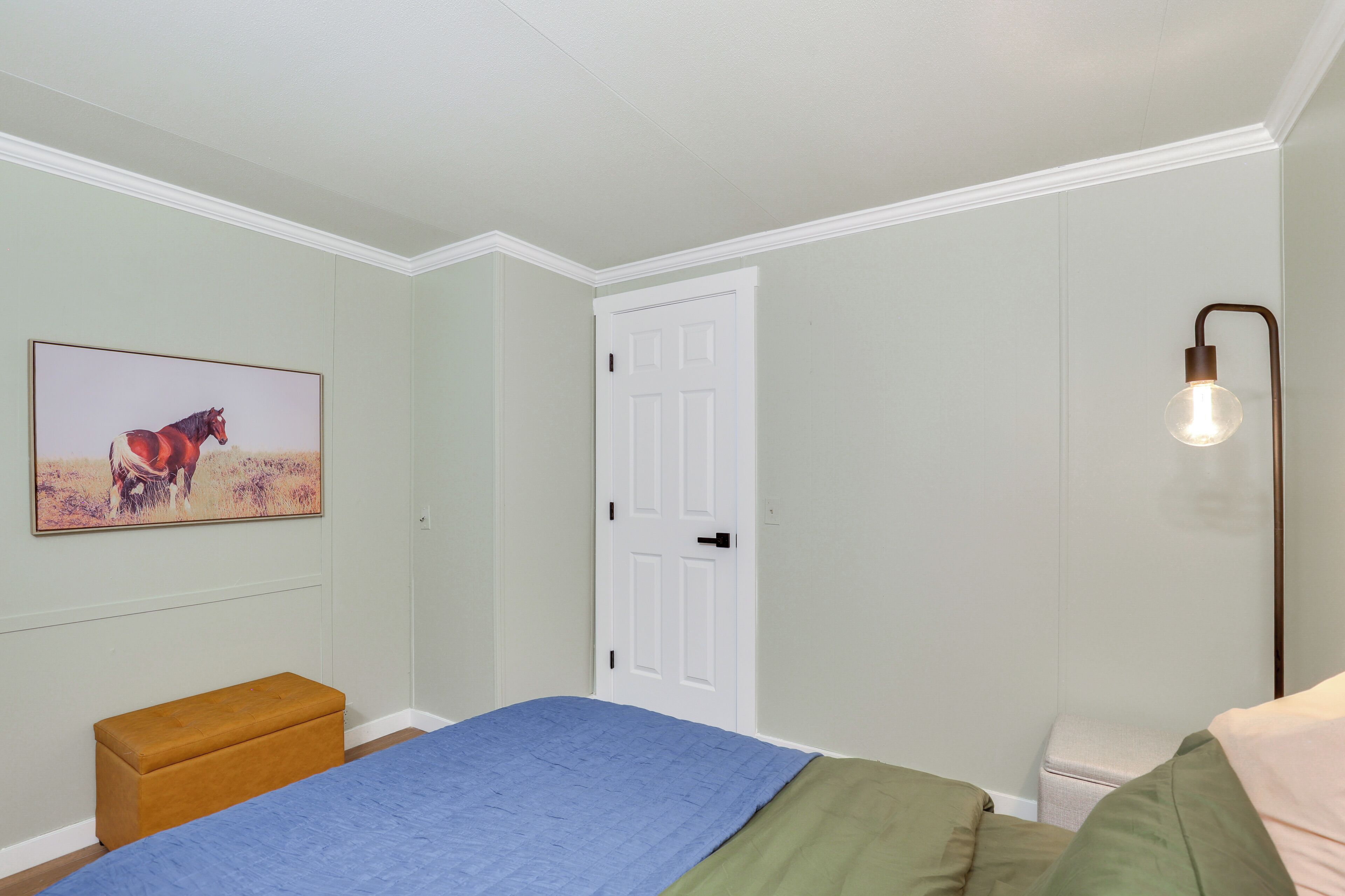 Um quarto com cama, mesa de cabeceira, banco, porta e luminária fixada na parede.