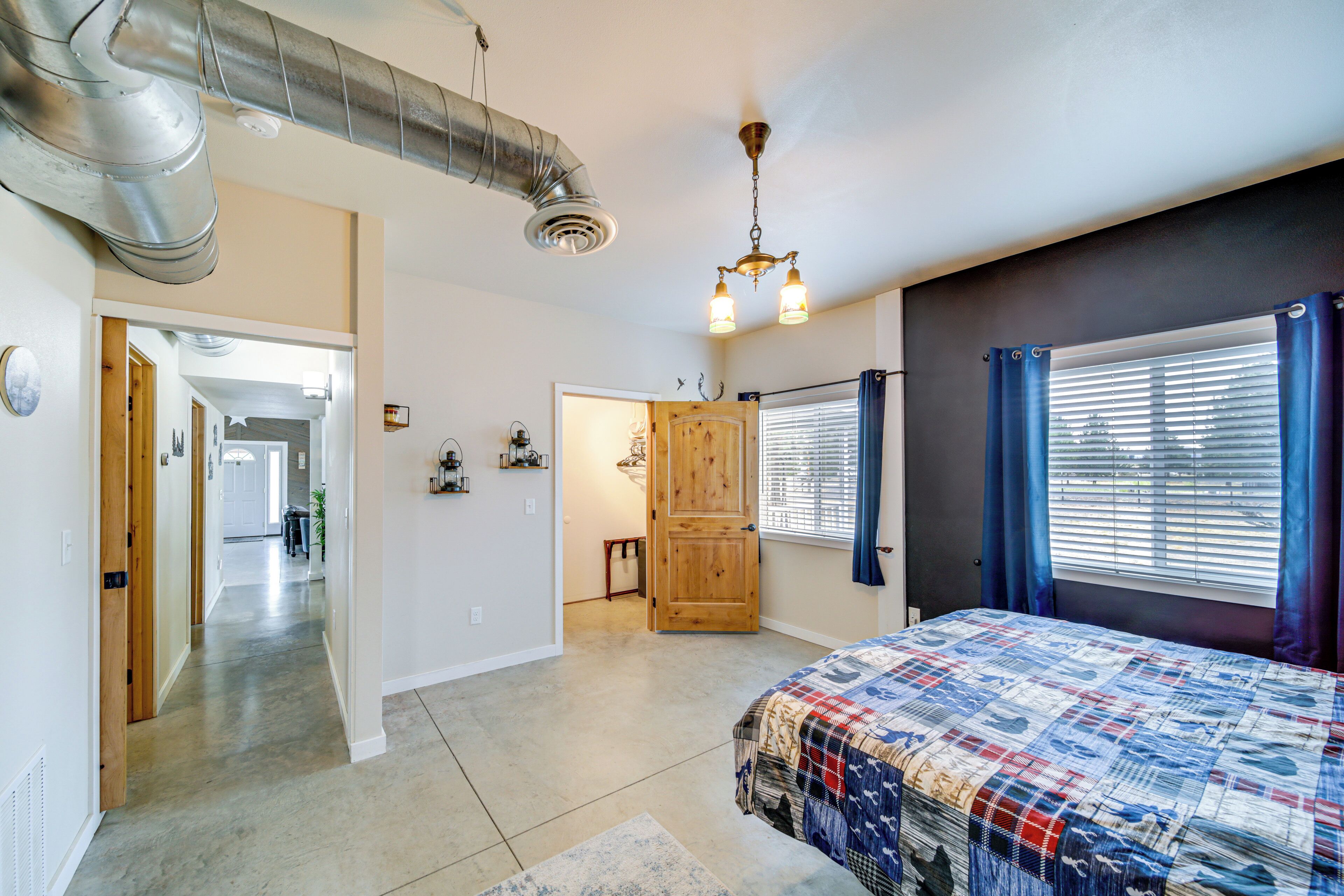 A bedroom with a large bed, a wooden door, a window with blinds, and a hallway leading to another room.