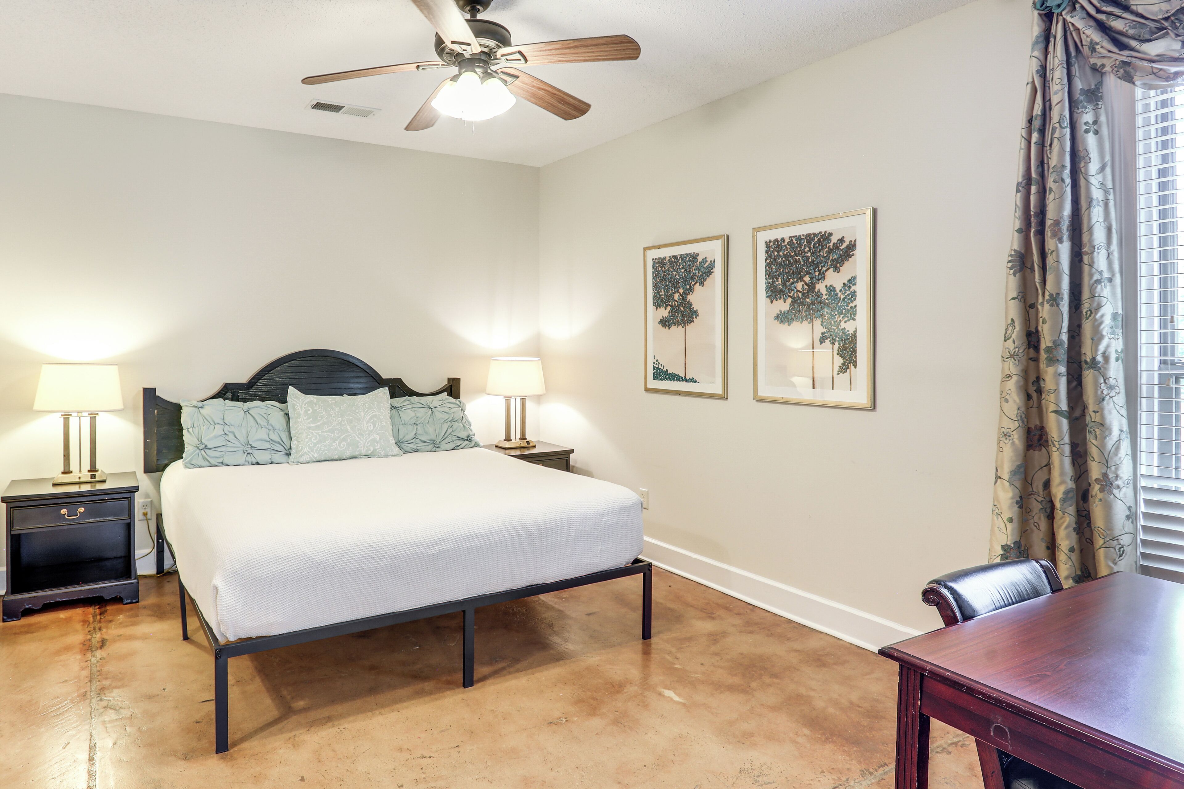Un dormitorio con cama, dos mesitas de noche con lámparas, un ventilador de techo y dos cuadros en la pared.