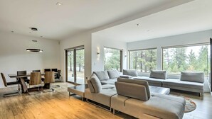 Ein modernes Wohnzimmer mit einem Ecksofa, einem Esstisch mit Stühlen und großen Fenstern mit Blick auf Bäume.