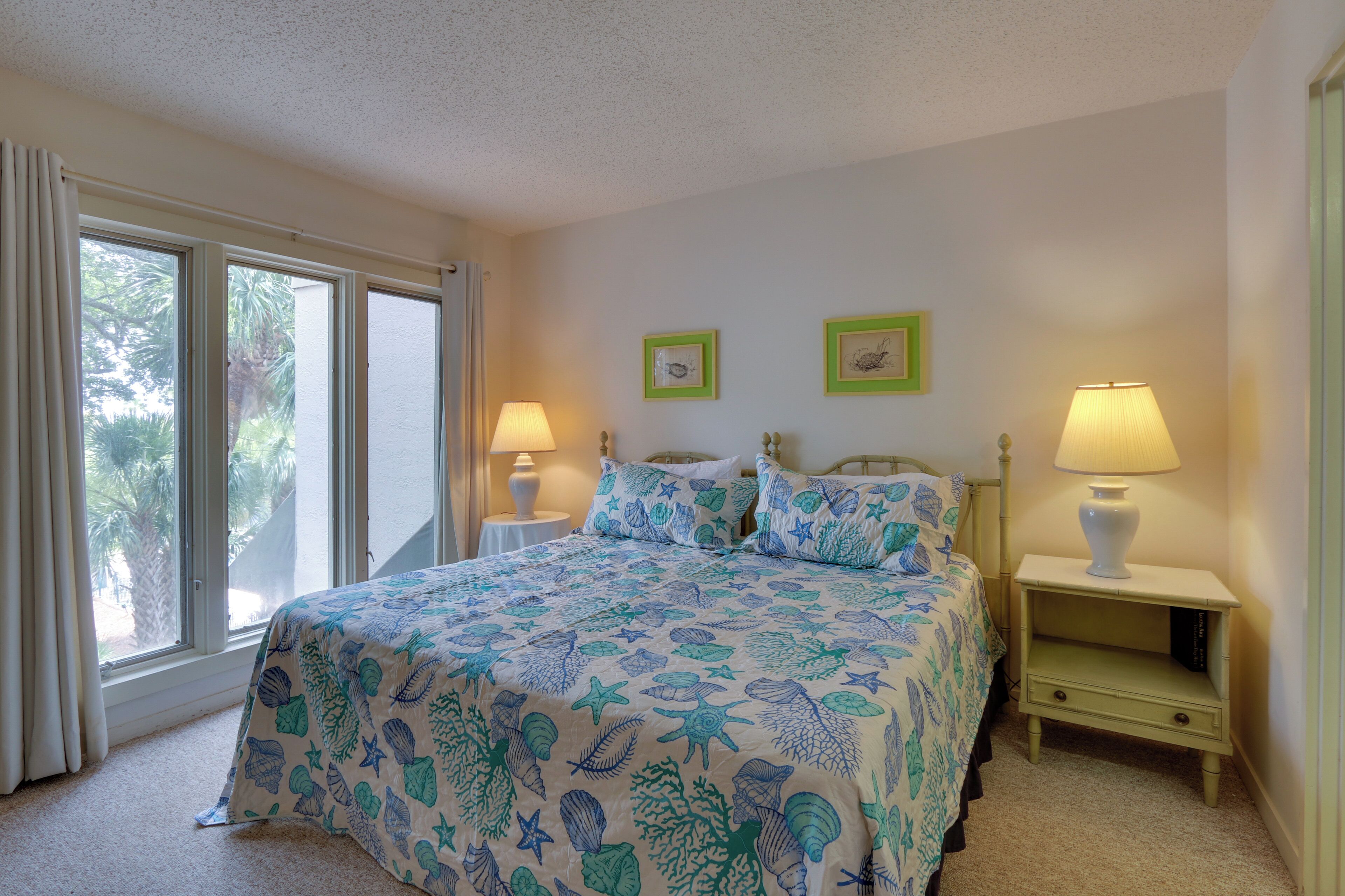 A bedroom with a bed, two bedside tables with lamps, a window with curtains, and two framed pictures on the wall.