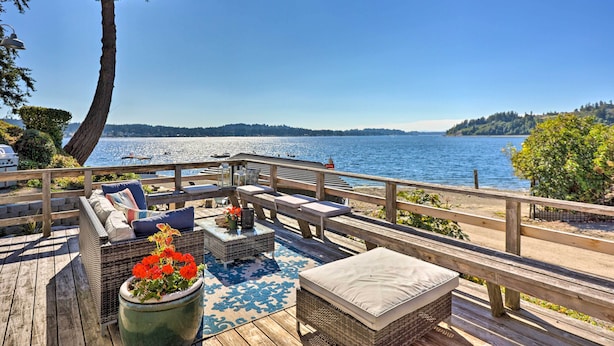 A wooden deck with outdoor furniture, a large planter with flowers, and a view of a lake and distant hills.