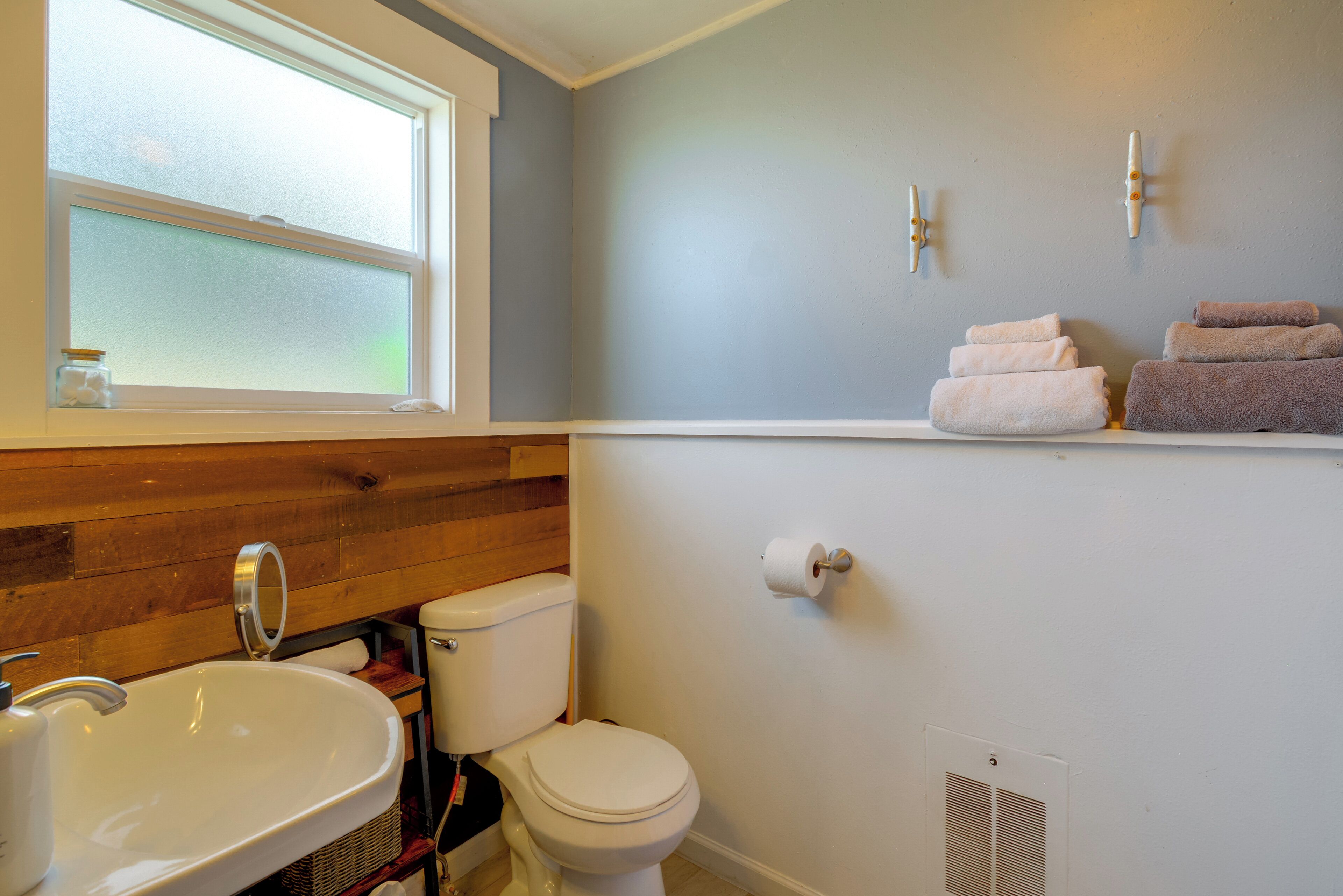 A bathroom with a wooden vanity, a white sink, a toilet, and towels on a shelf.