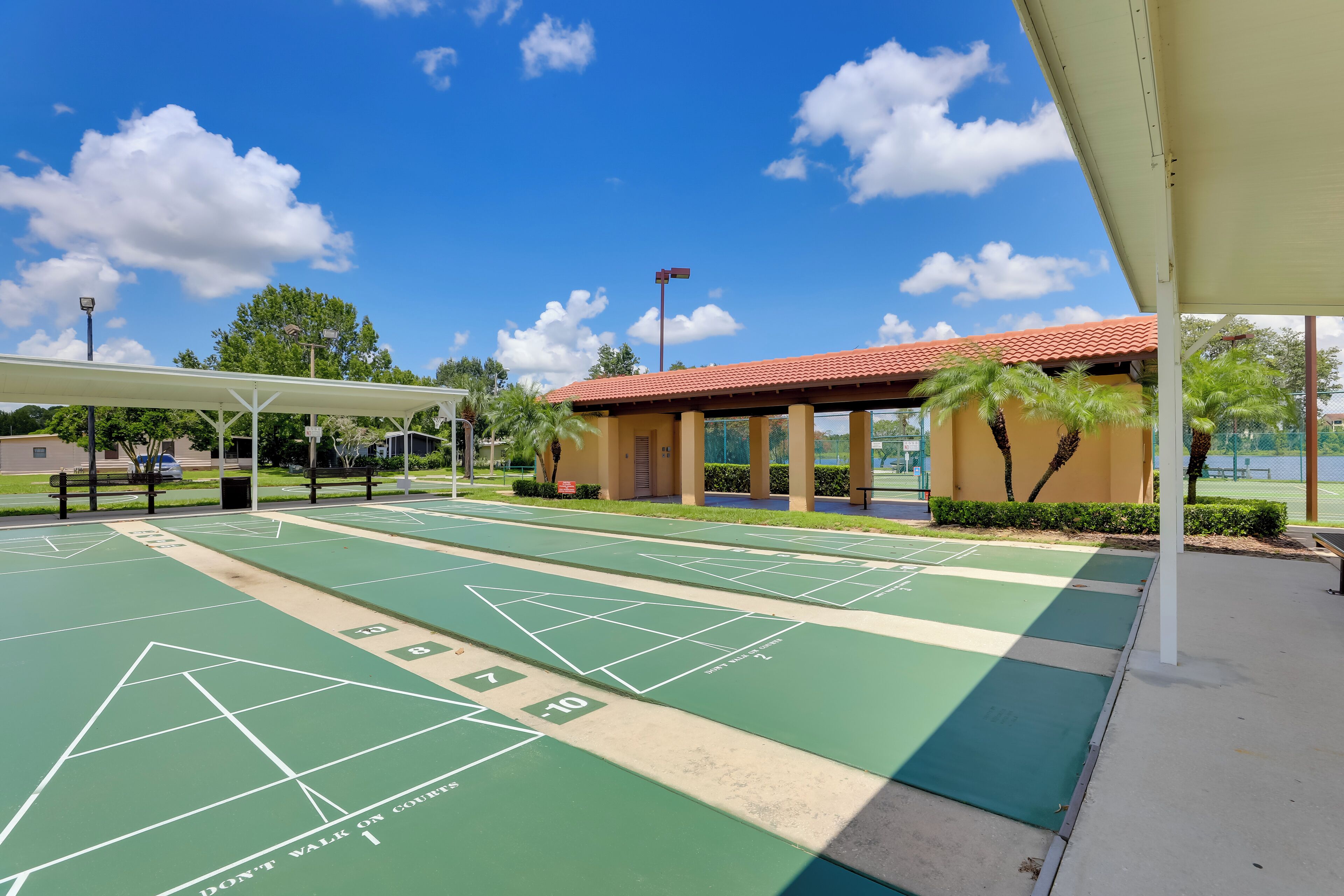 Ein Shuffleboard-Spielfeld mit grünlichen und beigen Markierungen, ein überdachter Sitzbereich und ein Gebäude mit rotem Ziegeldach.