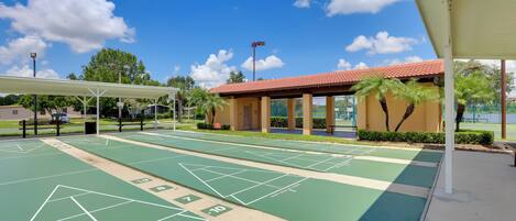 Ein Shuffleboard-Spielfeld mit grünlichen und beigen Markierungen, ein überdachter Sitzbereich und ein Gebäude mit rotem Ziegeldach.