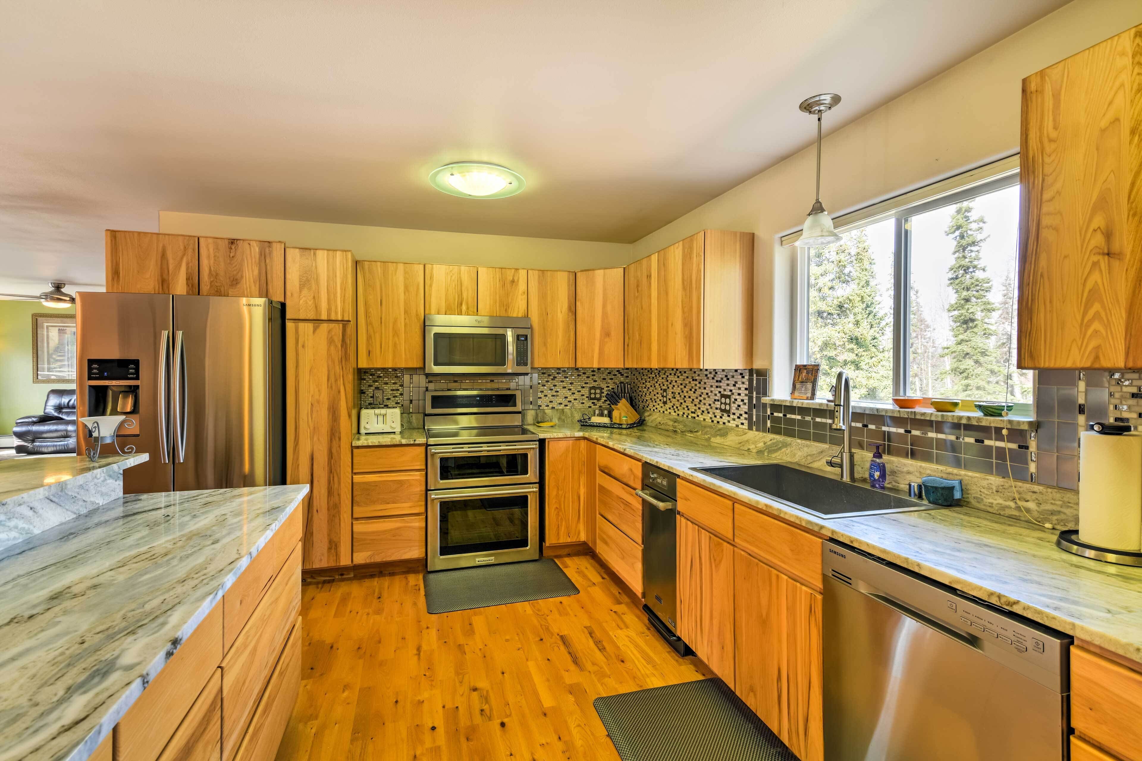 A modern kitchen with wooden cabinets, stainless steel appliances, a large window, and a marble countertop.
