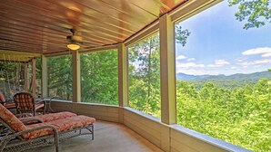 A covered porch with two wicker chairs and a table, overlooking a forested landscape. - Mountain View Home: Hike, Raft, Golf + More! (Bryson City)