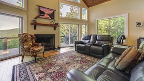A spacious living room with a fireplace, leather sofas, and large windows offering a view of the outdoors. - Mountain View Home: Hike, Raft, Golf + More! (Bryson City)