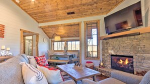 A cozy living room with a stone fireplace, a flat-screen TV mounted above it, and a wooden ceiling. - Pet-friendly Cabin w/ Mtn Views in Chinquapin! (Glenville)