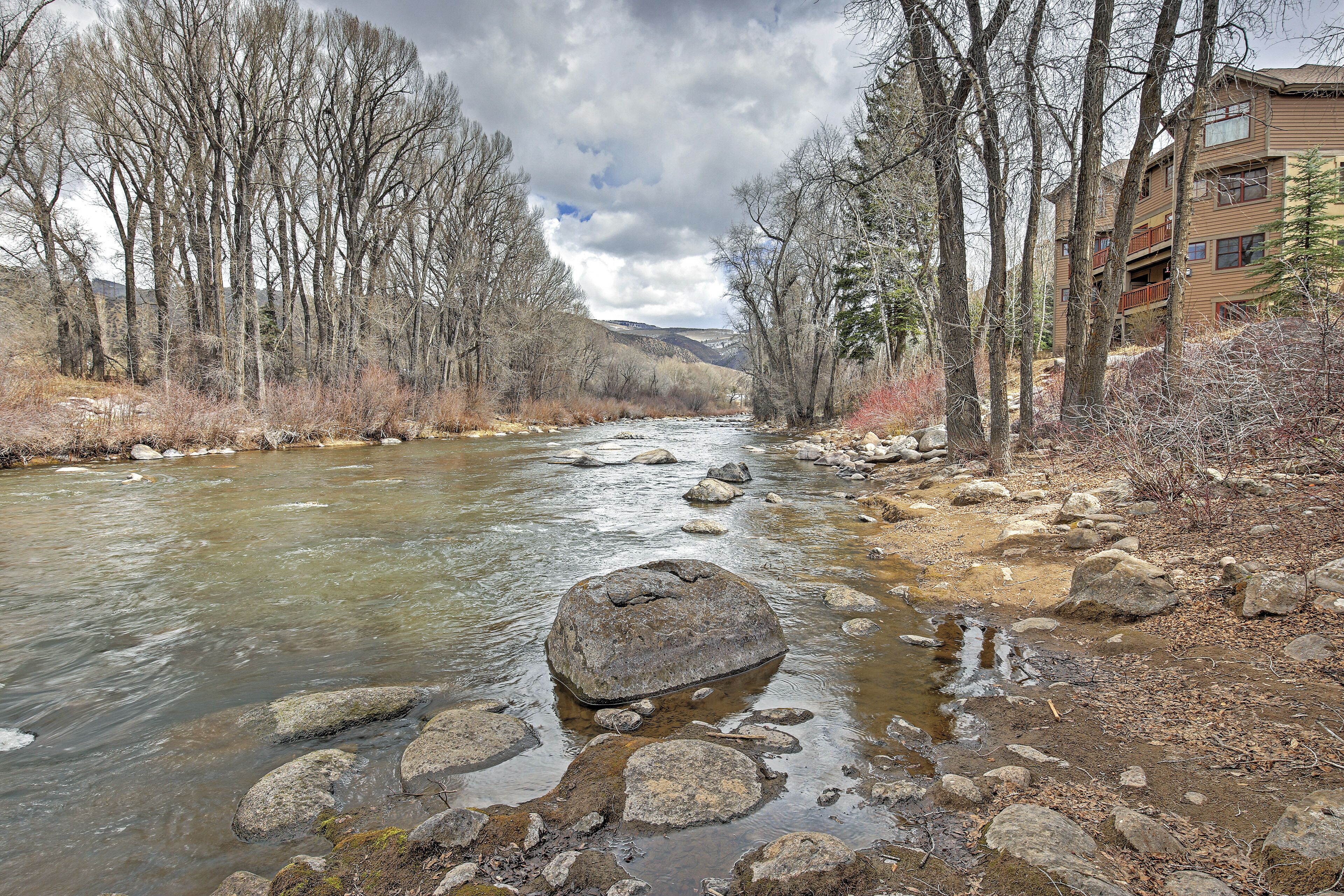 A river with rocks and trees, a building with balconies, and a mountainous background.