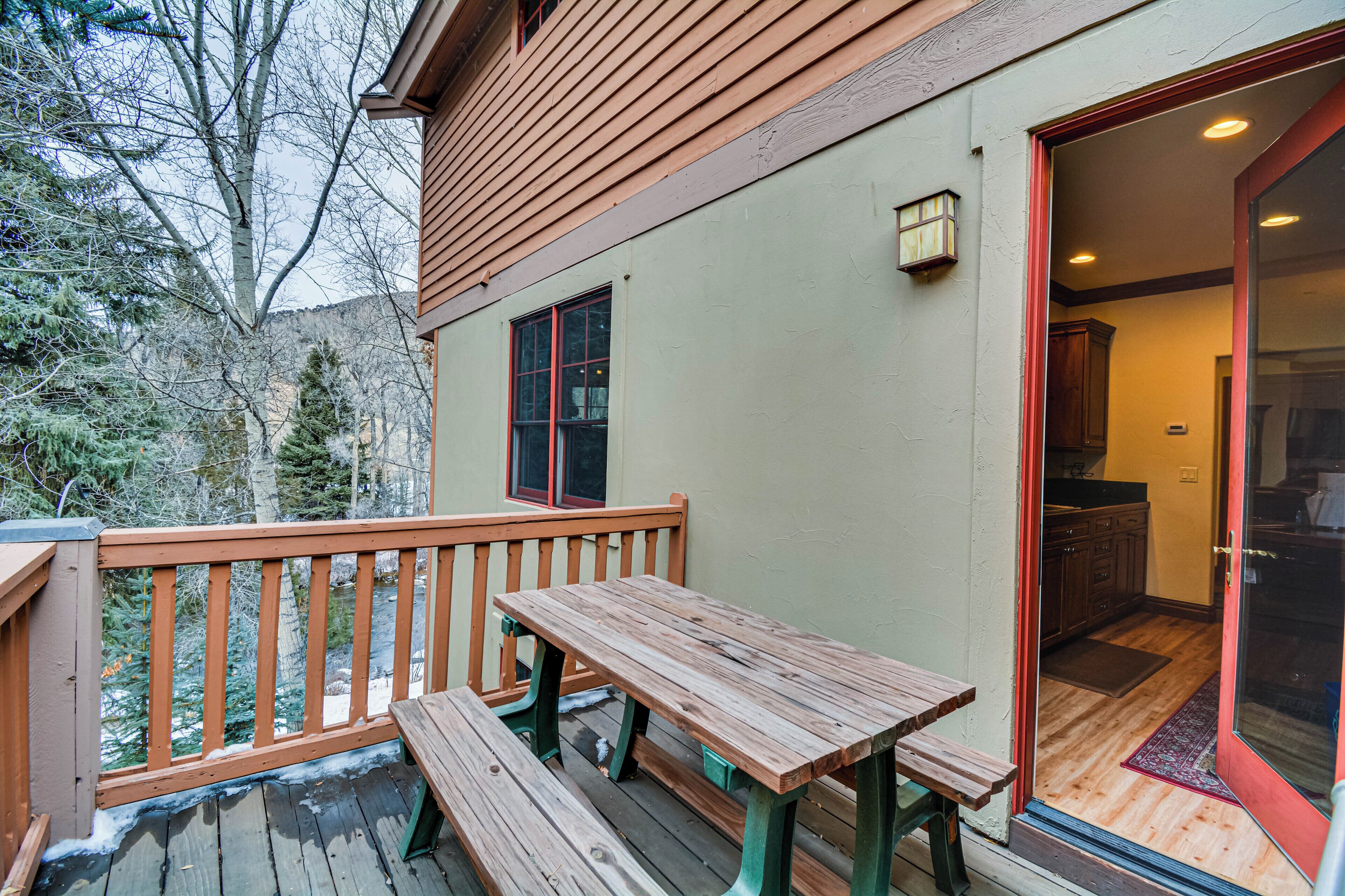 A wooden deck with a bench and a table, a door leading to a kitchen, and a view of trees and mountains in the background.