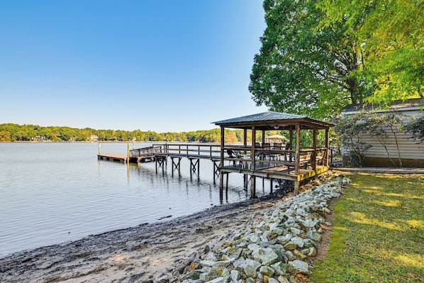 A lakeside gazebo with a wooden deck extending into the water, surrounded by a stone wall and a grassy area.