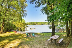 A lakeside picnic area with a wooden table and chairs, a canoe, and a dock extending into the water.