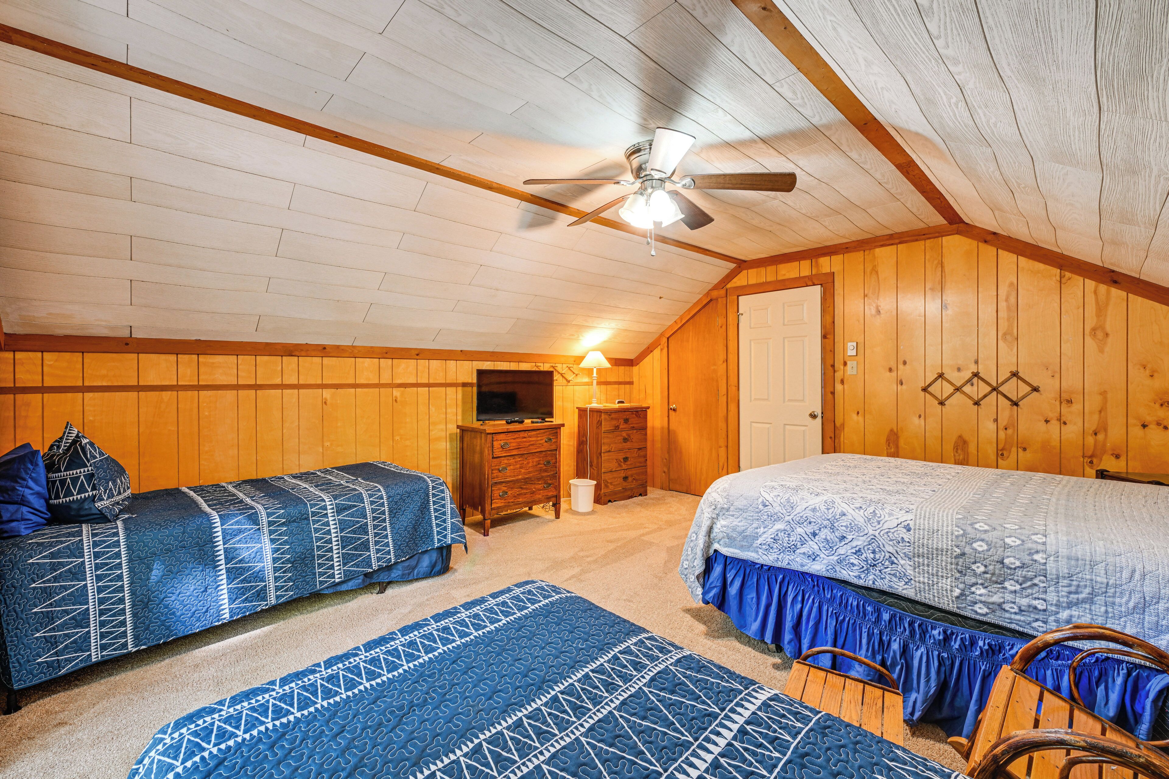 A cozy attic bedroom with two beds, a ceiling fan, wooden walls, and a television.