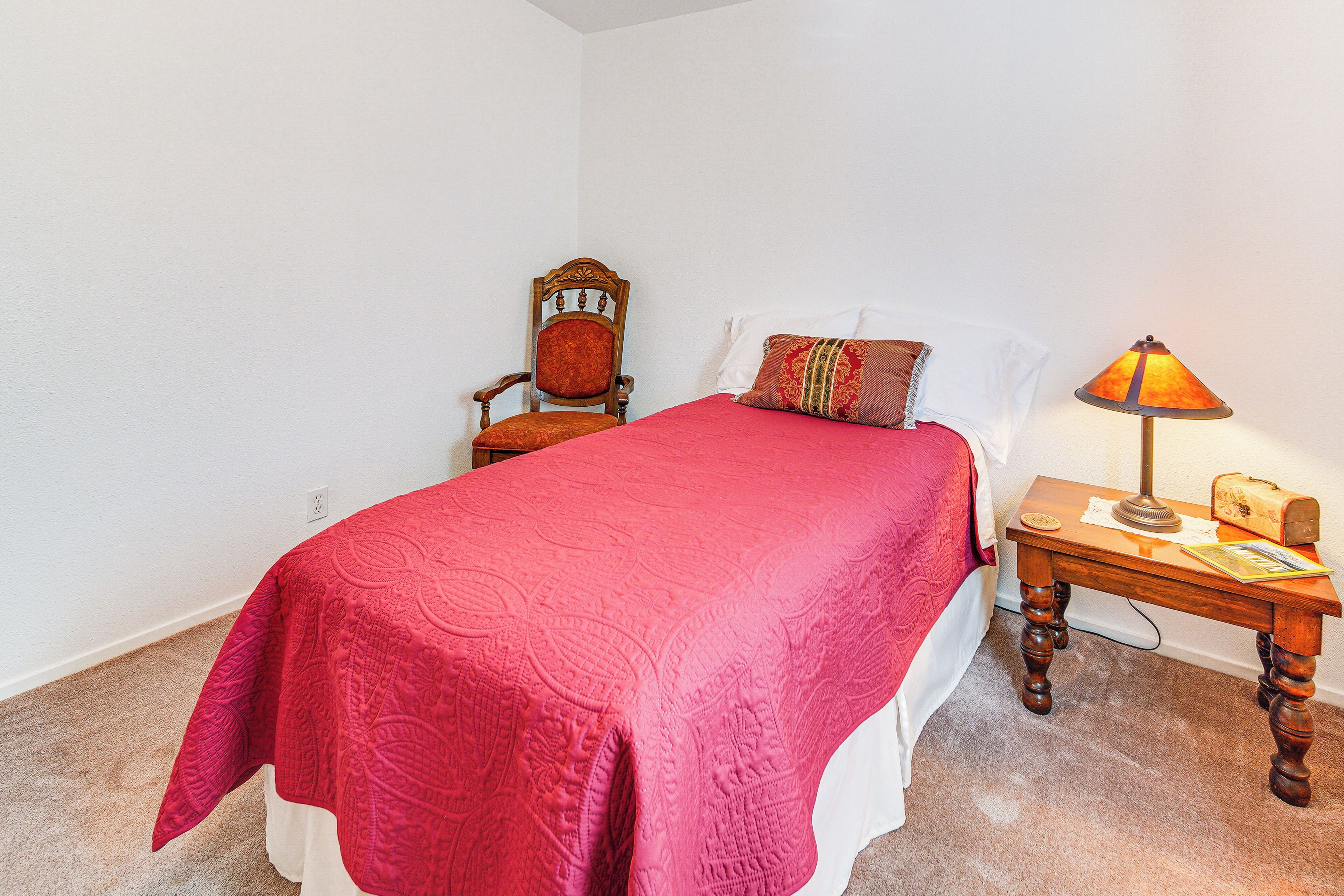 A bedroom with a bed, a wooden chair, a nightstand with a lamp, and a carpeted floor.