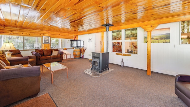 A cozy living room with wooden ceilings, a fireplace, and large windows offering views of the outdoors. - Mtn Gate Guest House ~ 6 Mi to Shasta Lake! (Redding)