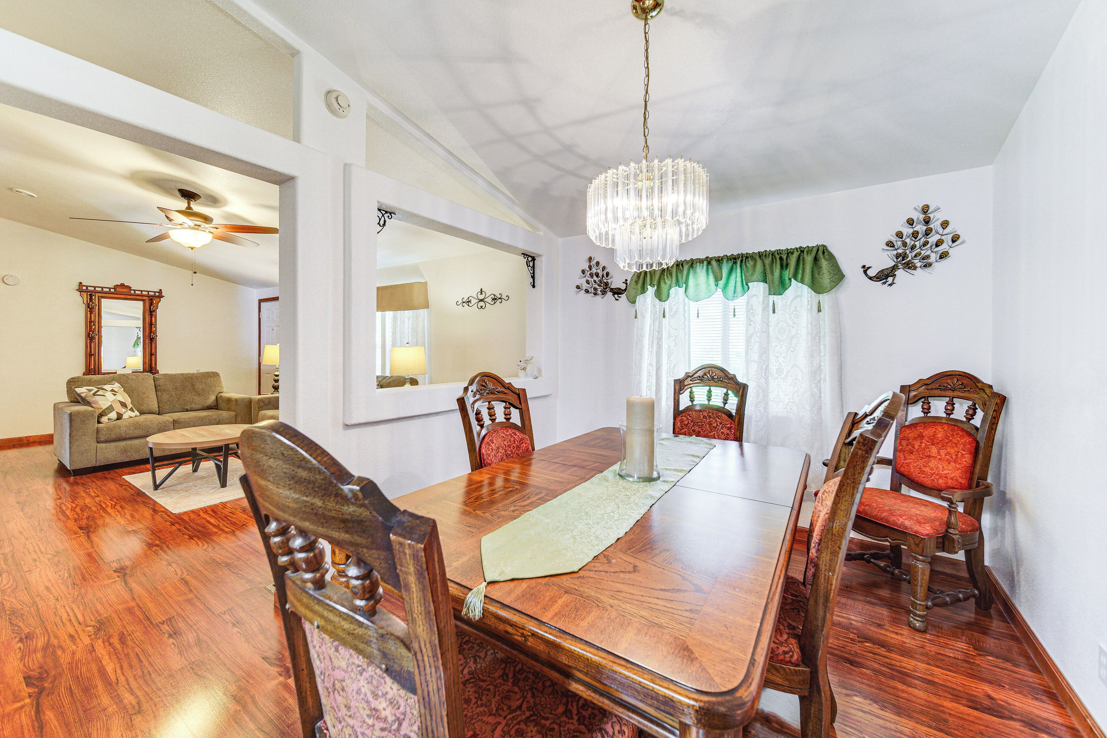 Un comedor con mesa de madera, sillas, una lámpara de araña y vista a una sala de estar con sofá y ventilador de techo.