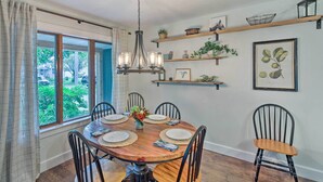 A dining room with a wooden table, six chairs, a chandelier, and a view of greenery through the window. - Airy Cottage Close to Rock Lake w/ Fire Pit! (Lake Mills)