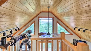 Un intérieur en bois agrémenté d’un télescope, d’un ventilateur de plafond et d’une grande fenêtre donnant sur la forêt.