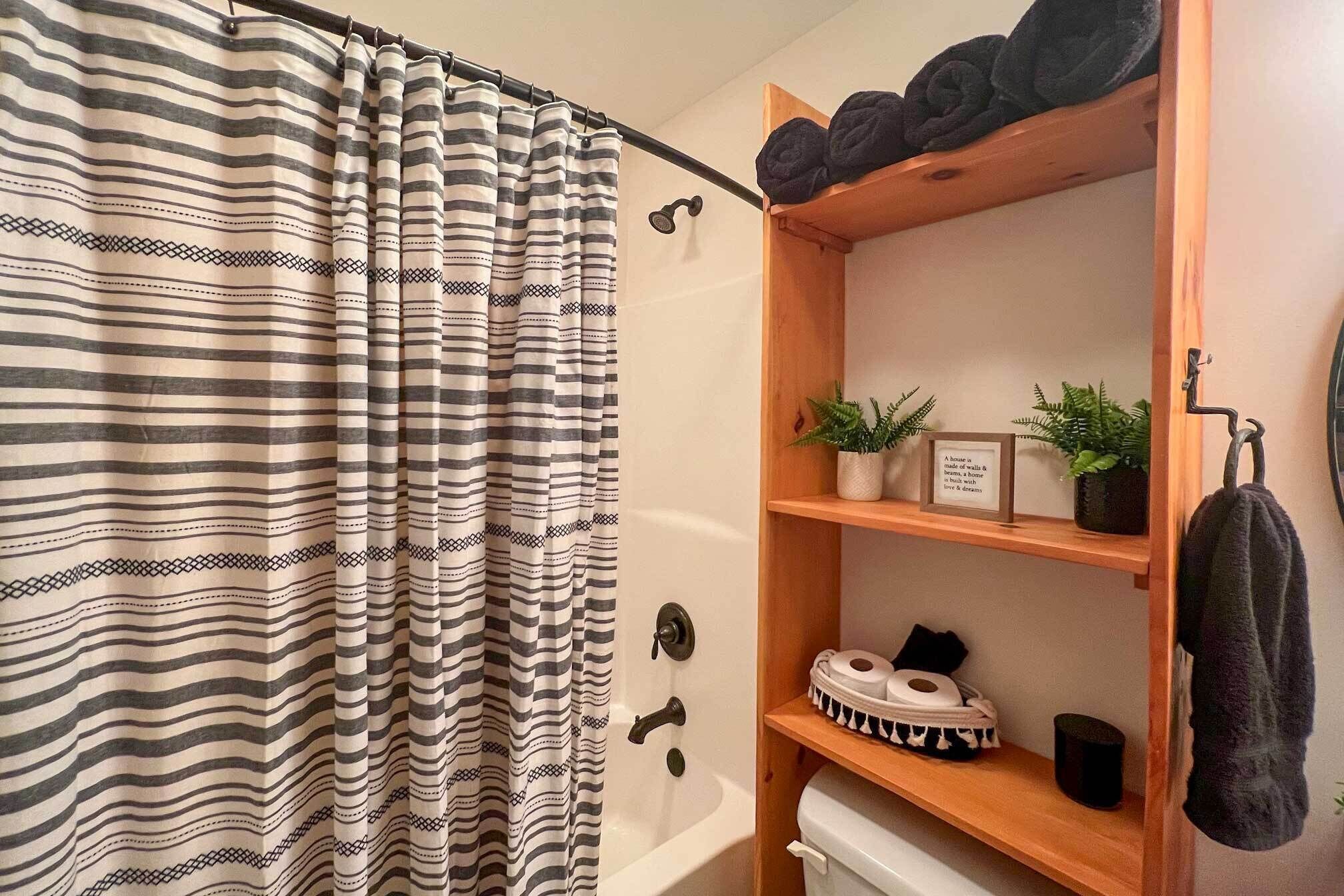 A bathroom with a striped shower curtain, a wooden shelf with plants and toiletries, and a towel hanging on a hook.