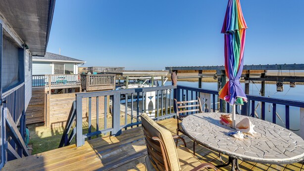 Eine Holzterrasse mit Tisch und Stühlen, einem bunten Sonnenschirm und Blick auf das Wasser.