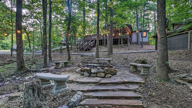 A rustic outdoor area with stone steps leading to a fire pit, surrounded by trees and a wooden deck with string lights. - Broken Bow Oasis: Hot Tub, Fire Pit & Patio (Broken Bow)