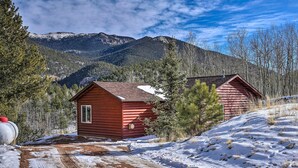 Une cabane accessible par un sentier de terre, entourée de neige et d’arbres, avec des montagnes en arrière-plan.