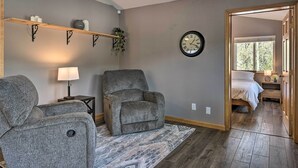 Un salon avec un fauteuil gris, une étagère en bois ornée d’objets décoratifs, une horloge murale et une vue sur une chambre à coucher, visible à travers une porte ouverte.