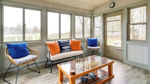 A sunlit room with a wooden ceiling, large windows, a sofa with blue and orange cushions, two wicker chairs, and a glass-topped coffee table. - Cadillac Vacation Rental w/ Dock on Lake Mitchell! (Cadillac)