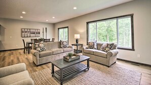 A spacious living room with a large window, a sofa set, a coffee table, and a dining area. - Upscale Townhome in Causeways on Gull Resort! (Nisswa)