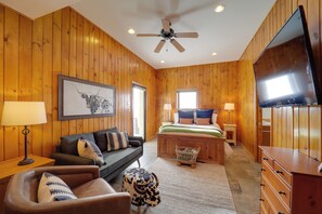 A room with wooden paneling, a ceiling fan, a bed with a green bedspread, a brown leather sofa, a small cow-shaped ottoman, and a flat-screen TV. - Views & Hot Tub! Beech Mtn 'sky Barn Retreat' (Banner Elk)