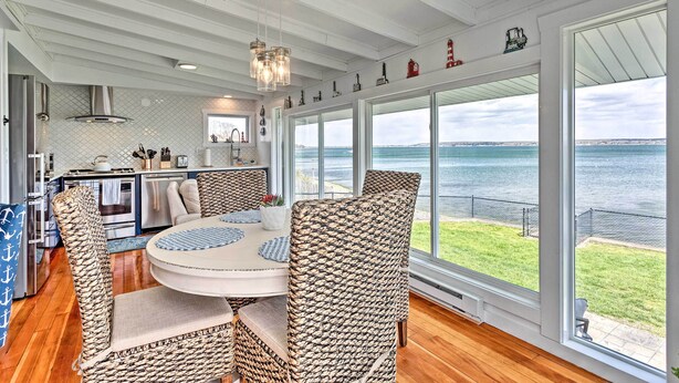 A dining area with wicker chairs and a round table, large windows with a view of the sea, and a kitchen with modern appliances. - Waterfront Portsmouth Cottage: 8 Mi. From Newport! (Portsmouth)