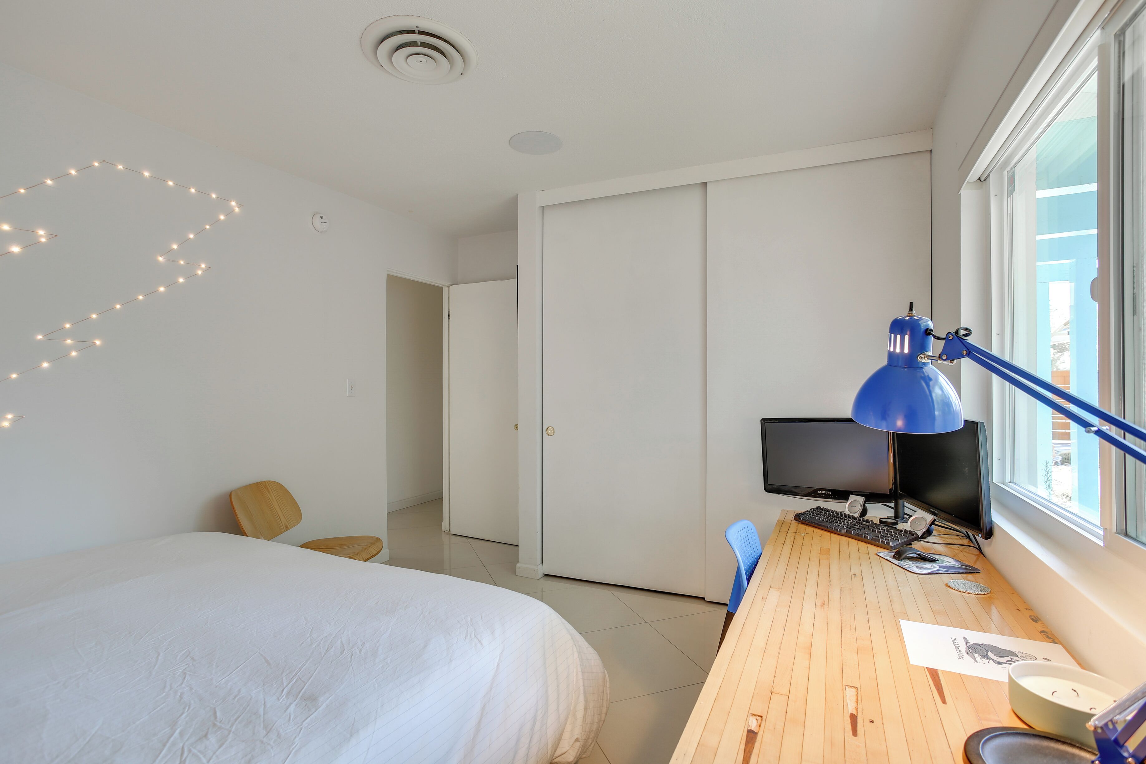 A bedroom with a bed, a desk with a computer, a blue desk lamp, and a window with a view.