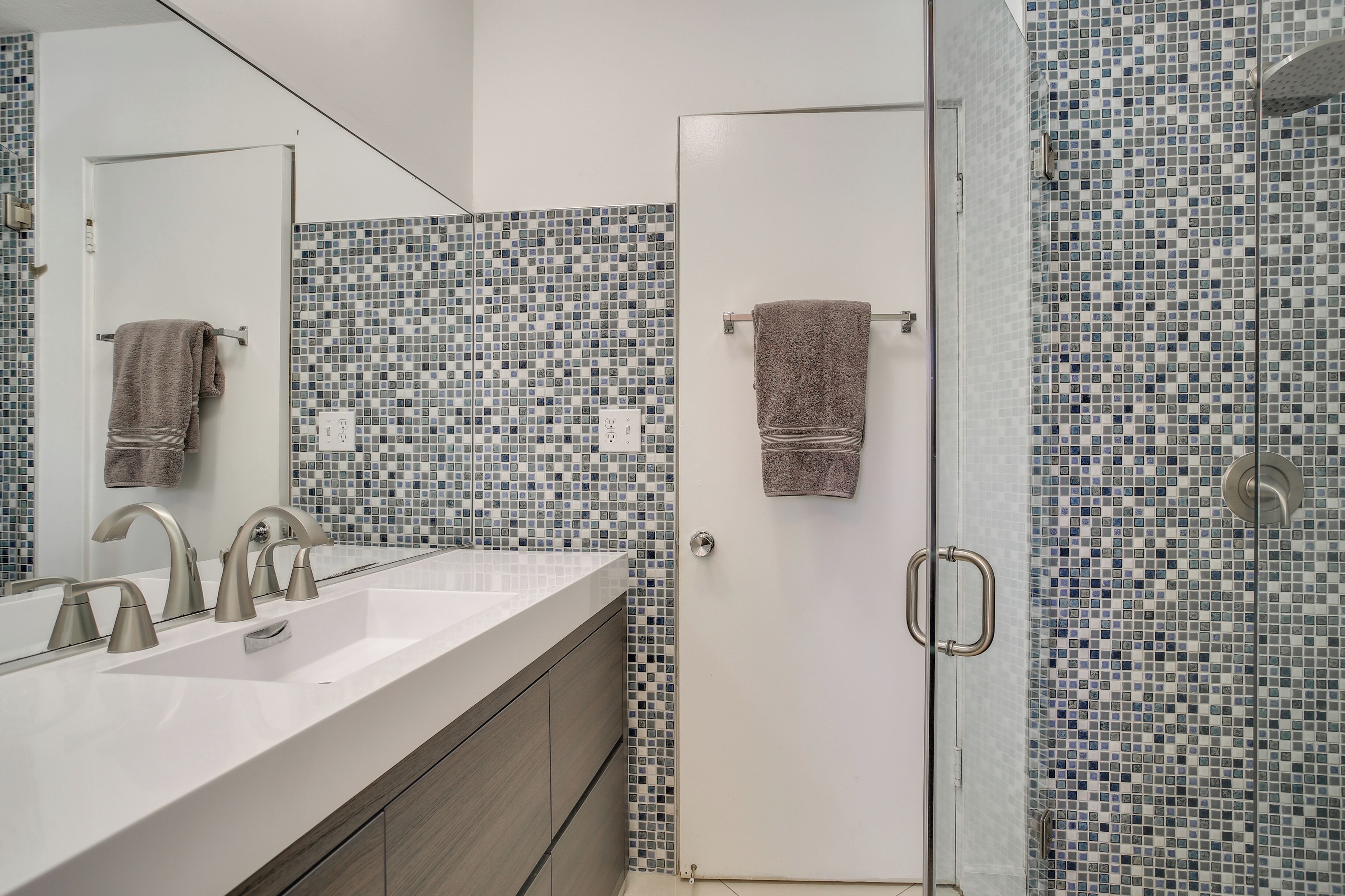 Een badkamer met een wit wastafel, twee kraan, een spiegel en een douche met mozaïek betegelde wanden.