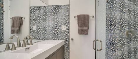 Een badkamer met een wit wastafel, twee kraan, een spiegel en een douche met mozaïek betegelde wanden.