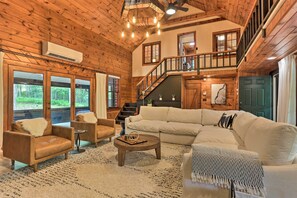 A spacious living room with a large sectional sofa, a coffee table, and two armchairs. There is a staircase leading to an upper level, and a large window offering a view of the outdoors. - Hilltop Lackawaxen Gem w/ River View & Fire Pit (Lackawaxen)
